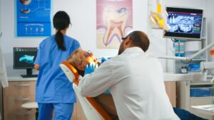 Dentist examining an older patient in a modern dental clinic with a dental assistant nearby.